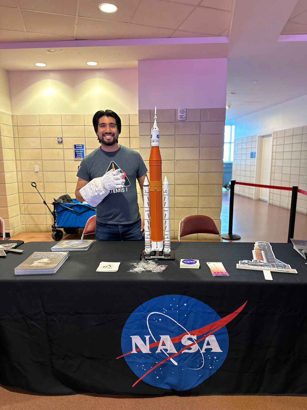 A NASA employee staffs an agency exhibit table with models and stickers at a community engagement event.